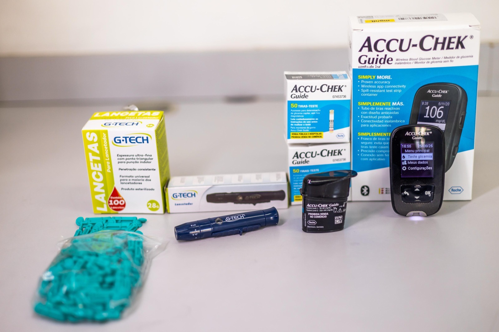 A imagem mostra um conjunto de itens utilizados para o controle de glicemia, organizados sobre uma superfície lisa e clara. São eles: Monitor de glicose Accu-Chek Guide – À direita da imagem, com visor ligado exibindo dados como horário (16:55), data (25/06/25) e nível de glicose de 106 mg/dL. O aparelho está em pé ao lado da embalagem maior, que tem o nome