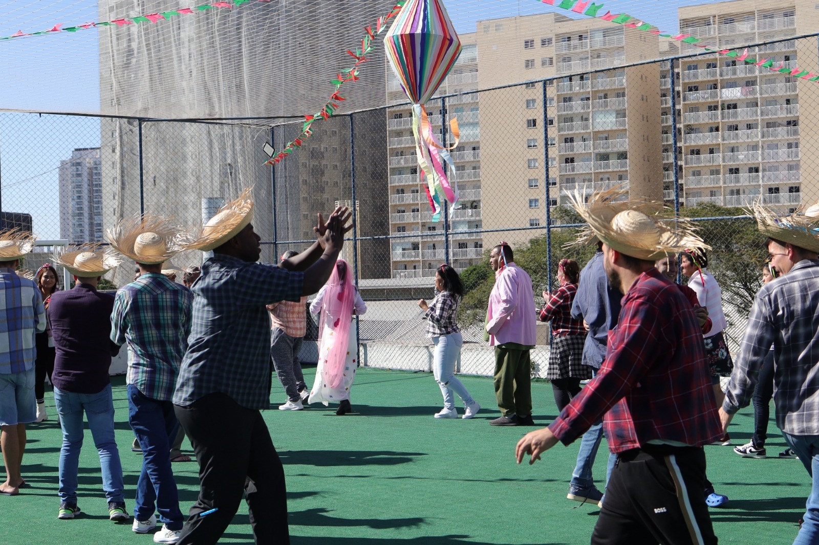 A imagem mostra um grupo de pessoas participando de uma festa junina ao ar livre, possivelmente em uma quadra esportiva de um prédio, cercada por telas de proteção e com prédios altos ao fundo. O clima é ensolarado. Homens e mulheres, adultos, estão vestidos com roupas típicas dessa celebração popular brasileira: camisas xadrez, calças jeans e chapéus de palha. Algumas mulheres usam vestidos e véus cor-de-rosa, em referência a noivas de quadrilha junina. Uma grande bandeirola colorida em forma de balão está pendurada no centro, e há bandeirinhas triangulares coloridas decorando o espaço. As pessoas estão em movimento, aparentemente dançando uma quadrilha ou brincando em roda. O ambiente transmite alegria, tradição e confraternização.