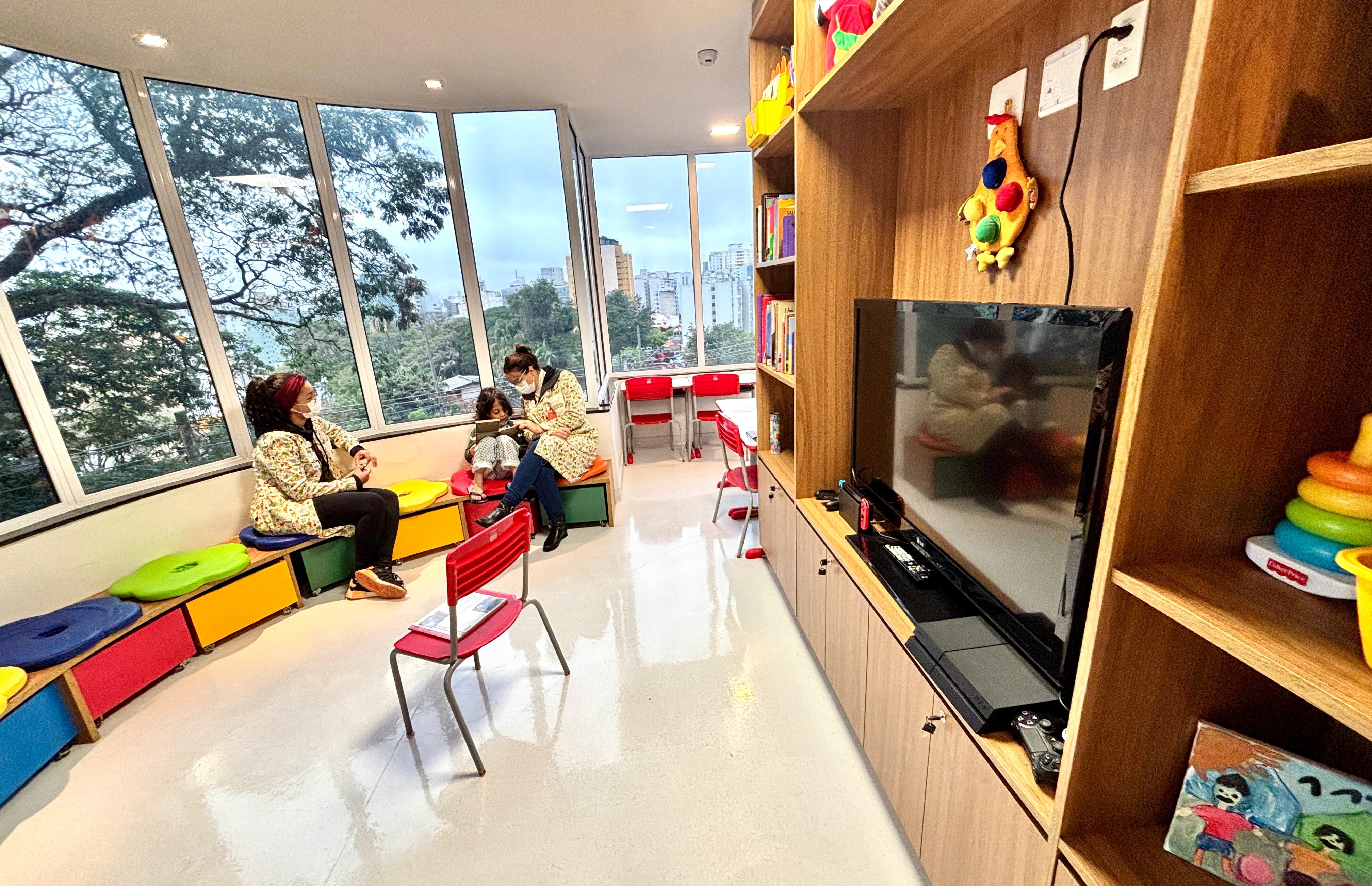 A imagem mostra um ambiente acolhedor e bem iluminado, provavelmente uma brinquedoteca ou sala de recreação infantil dentro de uma unidade de saúde ou hospital. O espaço tem janelas amplas que permitem a entrada de luz natural e oferecem vista para a cidade e a vegetação externa. À esquerda, duas mulheres usando jalecos coloridos (provavelmente profissionais da saúde ou voluntárias) interagem com duas crianças sentadas em bancos baixos e coloridos. Ao fundo, há mesas vermelhas infantis junto às janelas. À direita da imagem, vê-se uma estante de madeira com brinquedos, livros e uma televisão com um videogame instalado. Há também uma pequena cadeira infantil vermelha no centro do espaço. O ambiente é organizado, limpo e projetado para ser lúdico e confortável, promovendo bem-estar às crianças em atendimento.