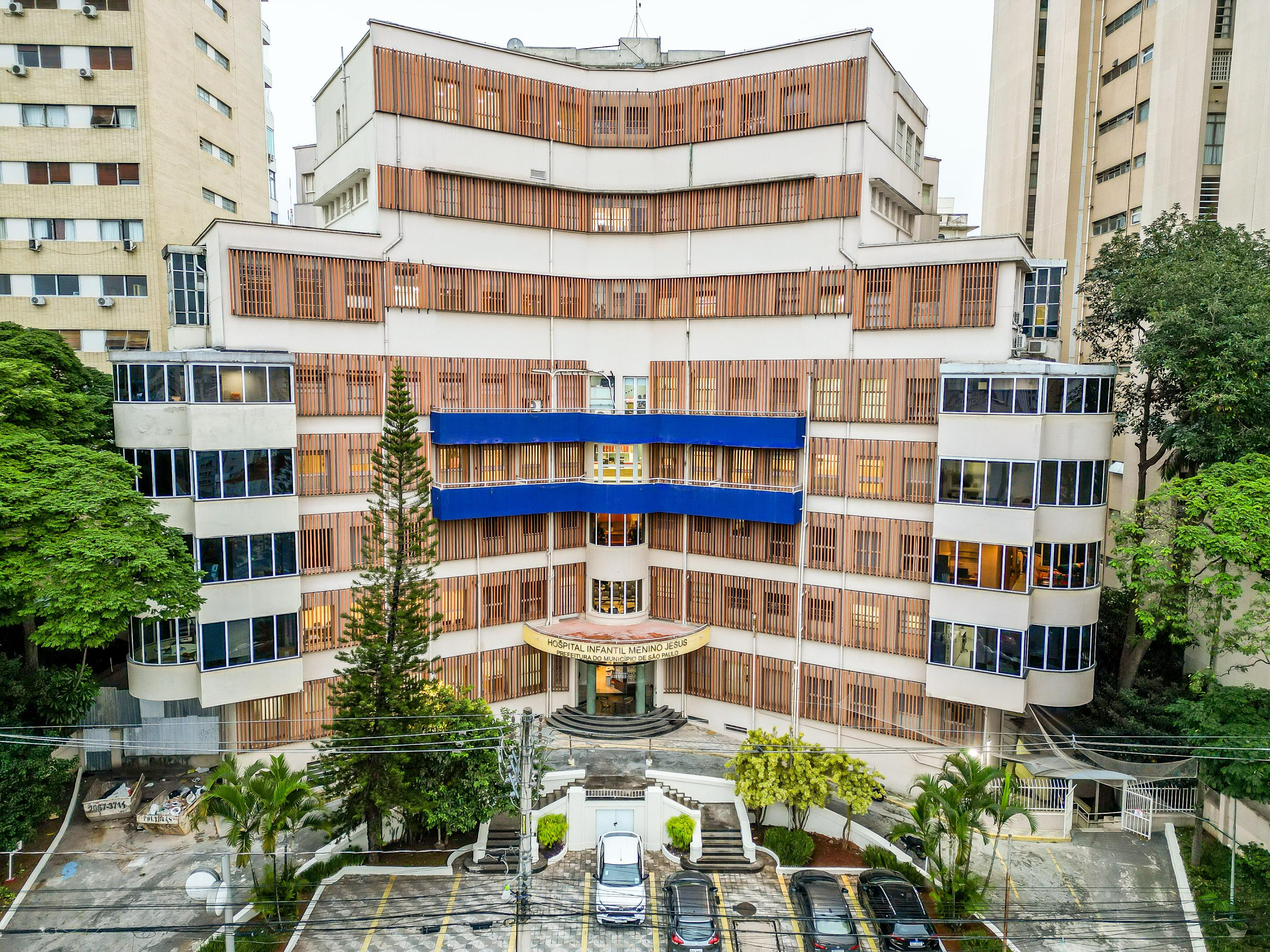 A imagem mostra a fachada do Hospital Infantil Menino Jesus, localizado em São Paulo. O prédio é de arquitetura moderna e simétrica, com várias janelas e estruturas verticais de madeira que funcionam como brises. Na parte central do edifício, destacam-se duas faixas horizontais azuis que percorrem os andares médios da construção.  O letreiro acima da entrada principal identifica claramente o local como “Hospital Infantil Menino Jesus”. Há uma escadaria semicircular que leva até a porta principal, ladeada por canteiros com vegetação. Em frente ao prédio, vê-se um pequeno estacionamento com carros estacionados e fios de eletricidade atravessando a imagem. O hospital está cercado por outros edifícios altos e árvores, compondo um ambiente urbano e arborizado.