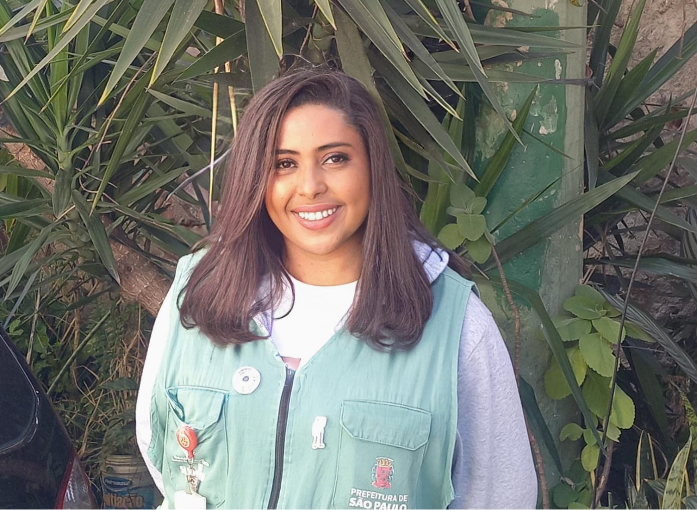 A imagem mostra uma mulher sorridente posando ao ar livre, em frente a uma vegetação com folhas grandes e verdes. Ela veste um colete verde com o logotipo da Prefeitura de São Paulo, indicando que está em serviço ou envolvida em alguma atividade institucional. O colete tem alguns bottons e um crachá pendurado. A mulher tem cabelos castanhos escuros e lisos, na altura dos ombros, e usa uma blusa clara com capuz por baixo do colete. A imagem transmite uma atmosfera positiva e acolhedora.