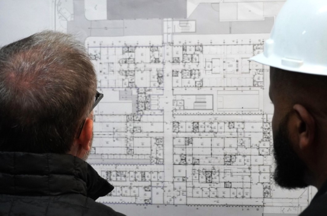 A imagem mostra dois homens observando atentamente a planta arquitetônica de uma construção. Um deles está usando capacete branco, indicando que provavelmente é um engenheiro ou técnico de segurança em uma obra. O outro homem, de cabelos grisalhos e óculos, veste um casaco escuro e também demonstra estar envolvido na análise técnica do projeto. A planta exibida contém diversos compartimentos, corredores e anotações técnicas, sugerindo um projeto de grande porte, possivelmente de um edifício público, hospitalar ou educacional. A cena retrata um momento de inspeção ou planejamento técnico, evidenciando o cuidado e o detalhamento envolvidos nas etapas de construção civil ou reforma de uma edificação.