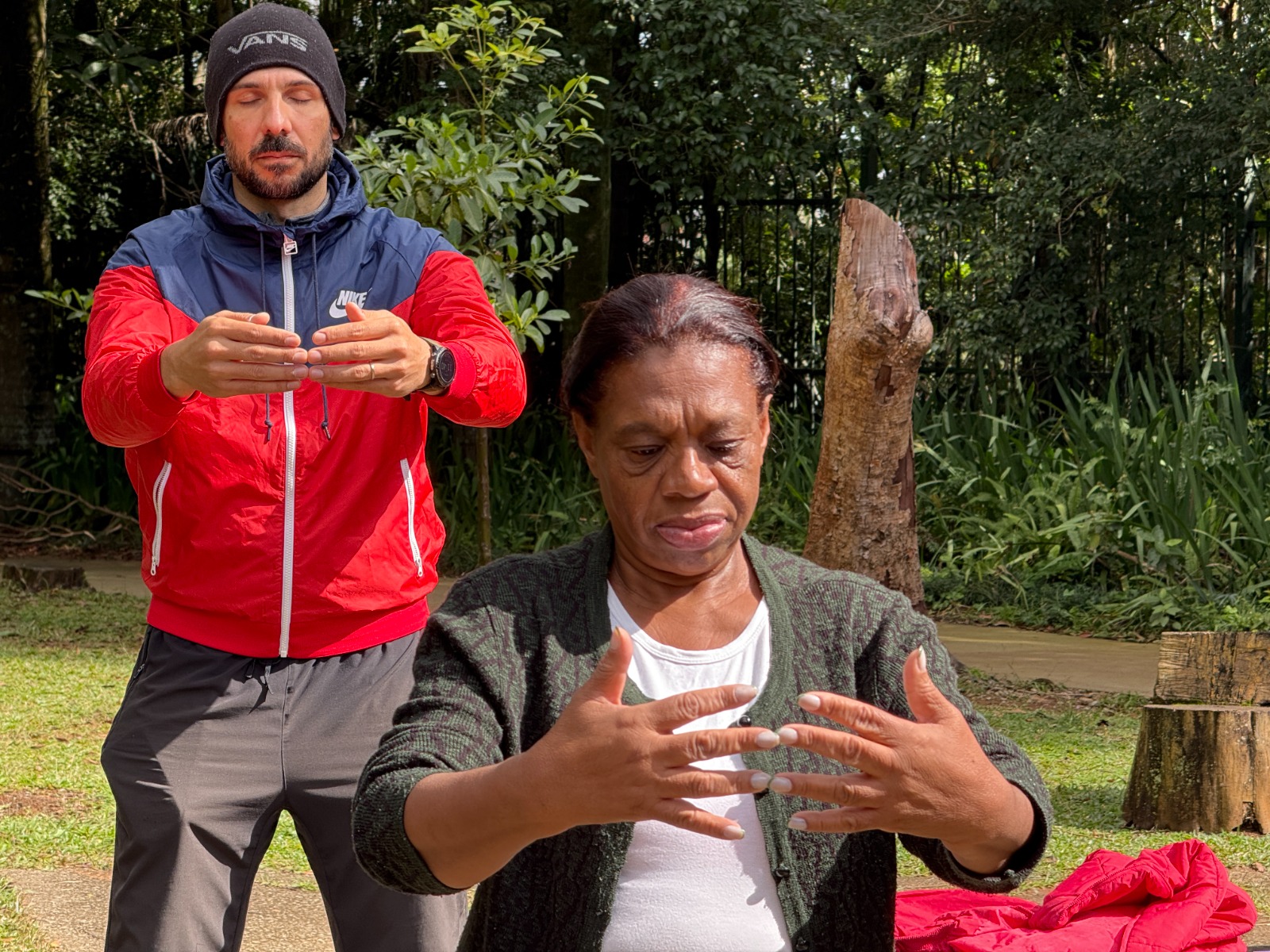 A fotografia mostra duas pessoas em um momento de concentração e conexão corporal, possivelmente praticando tai chi pai lin ao ar livre, em um ambiente arborizado. Em primeiro plano, está uma mulher negra de pele escura e cabelos presos para trás. Ela veste uma camiseta branca por baixo de um cardigã verde escuro. Seus olhos estão fechados e a expressão é séria e introspectiva. As mãos estão levantadas à frente do corpo, com os dedos suavemente curvados, como se ela estivesse sentindo a energia entre as palmas. Atrás dela, está um homem branco de barba, vestindo um agasalho da Nike vermelho com detalhes em azul-marinho e branco, calça cinza escura e gorro preto da marca Vans. Ele também está com os olhos fechados e as mãos na mesma posição que a mulher à frente, em um gesto calmo e deliberado, como se imitasse ou acompanhasse seus movimentos. O ambiente é natural, com grama, árvores, um toco de madeira ao lado e vegetação densa ao fundo. A luz do sol bate suavemente nas pessoas, especialmente iluminando o rosto da mulher. Há também uma jaqueta vermelha dobrada no chão à direita da imagem.