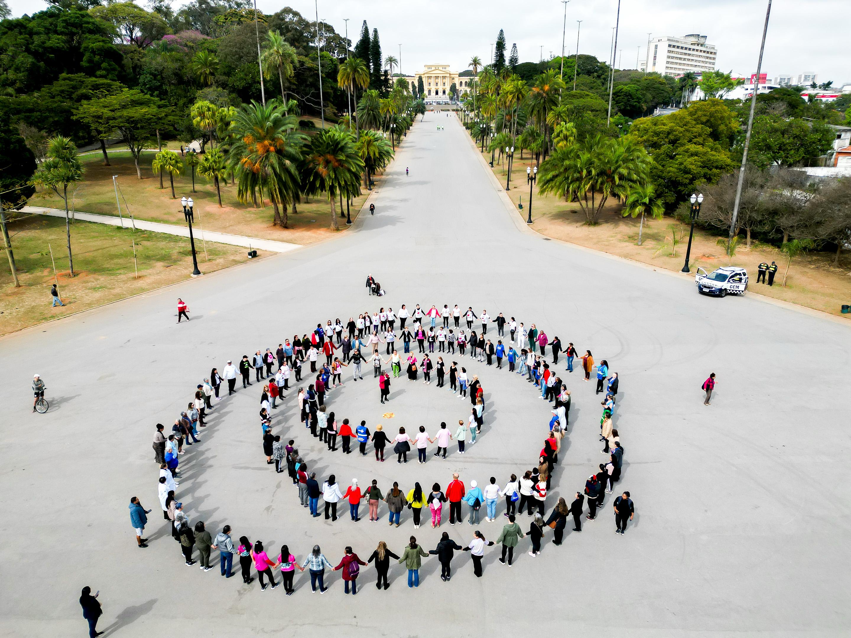 A imagem mostra uma grande formação humana em espiral, composta por dezenas de pessoas de mãos dadas, ocupando o centro de uma ampla avenida pavimentada. A espiral é formada por várias voltas concêntricas, com os participantes organizados de maneira bastante simétrica e coordenada. O local é o Parque da Independência, em São Paulo, identificado ao fundo pelo edifício imponente do Museu do Ipiranga, com sua arquitetura clássica e coloração bege clara. O museu está centralizado na composição da foto, ao fim da longa alameda arborizada que leva até ele. Há palmeiras imperiais e outras árvores ao longo dos dois lados da via. As pessoas que formam a espiral vestem roupas variadas, coloridas e casuais, indicando clima ameno. Há diversidade de idades, com predominância de adultos e idosos. A perspectiva aérea, feita provavelmente por um drone, ressalta a simetria da espiral humana e a imponência do espaço urbano e paisagístico. A cena transmite união, mobilização e propósito coletivo — possivelmente um ato simbólico ou manifestação pacífica.