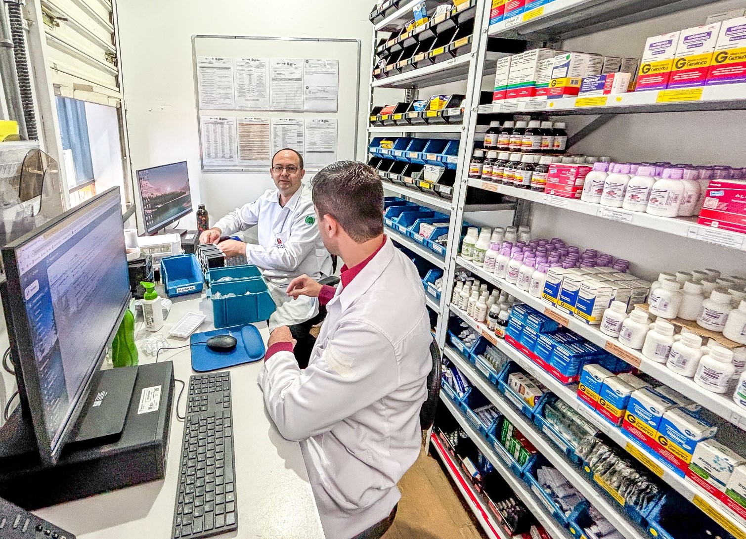 A imagem mostra dois profissionais de saúde, possivelmente farmacêuticos ou técnicos de farmácia, trabalhando em uma farmácia hospitalar ou unidade de dispensação de medicamentos. Ambos vestem jalecos brancos e estão sentados em frente a computadores, manuseando caixas e frascos de medicamentos.  À direita da imagem, há prateleiras organizadas com uma grande variedade de remédios — frascos, cartelas e caixas — muitos deles identificados como genéricos. No fundo, há uma parede com documentos afixados, provavelmente instruções, protocolos ou tabelas de controle. O ambiente é limpo, organizado e bem iluminado, sugerindo um espaço preparado para garantir segurança e eficiência no atendimento farmacêutico.
