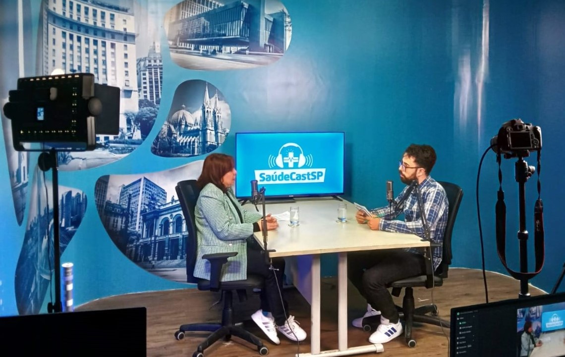 A imagem mostra a gravação de um podcast ou programa de entrevista chamado “SaúdeCastSP”.  Dois apresentadores/entrevistados estão sentados frente a frente em uma mesa branca, cada um com um microfone. Há copos de água sobre a mesa e um monitor ao fundo exibindo o logotipo do programa, que traz ícones de saúde e fones de ouvido.  O ambiente é um estúdio, com paredes azuis decoradas com imagens de pontos arquitetônicos de São Paulo. É possível ver equipamentos profissionais, como câmeras e iluminação, indicando que a entrevista está sendo registrada em áudio e vídeo.