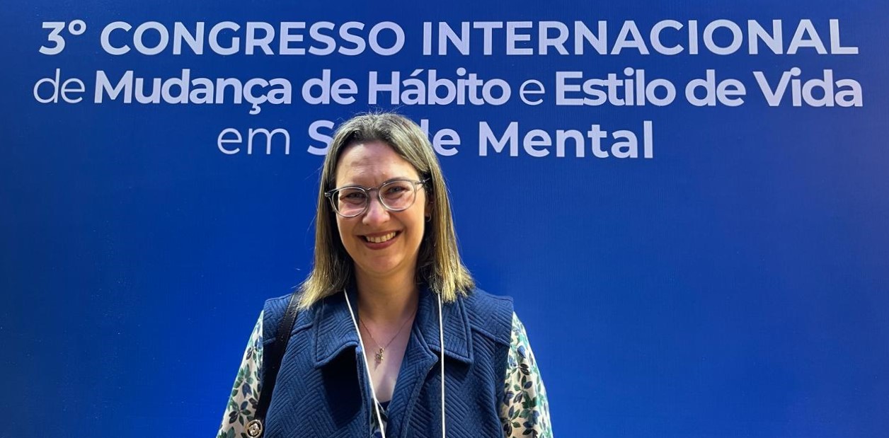 A imagem mostra Vanessa Junger sorrindo em frente a um painel azul que traz o texto: “3º Congresso Internacional de Mudança de Hábito e Estilo de Vida em Saúde Mental”. Ela tem pele clara, cabelo liso castanho-claro na altura dos ombros e usa óculos de armação transparente. Está vestindo uma blusa com estampa floral em tons claros e um colete azul marinho acolchoado. Usa um cordão com crachá no pescoço e carrega uma bolsa preta no ombro esquerdo. O fundo é totalmente azul, destacando o texto em branco e azul-claro.