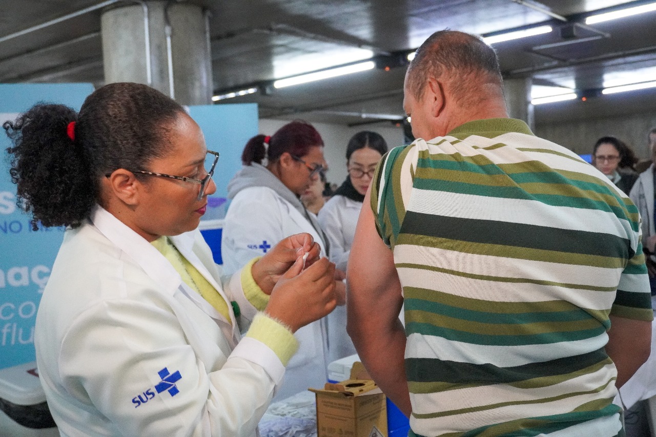 A imagem mostra uma profissional de saúde, vestindo jaleco branco com o logotipo do SUS no braço, aplicando uma vacina no braço esquerdo de um homem que veste uma camiseta listrada em tons de verde, branco e amarelo. A cena ocorre em um ambiente interno, possivelmente um posto de vacinação provisório, com outras profissionais de saúde ao fundo, também de jaleco, preparando materiais. A iluminação é artificial, com lâmpadas no teto, e há uma estrutura de concreto visível