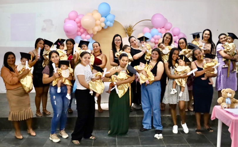 A imagem mostra um grupo de mulheres reunidas em um palco, cada uma segurando um bebê vestido com beca dourada e chapéu de formatura. Algumas crianças também seguram diplomas simbólicos. Ao fundo, há uma decoração com arco de balões nas cores rosa, azul e dourado, com um painel circular central. As mulheres e bebês estão sorrindo, sugerindo um clima de celebração, possivelmente de um evento de formatura simbólica para os bebês. À direita, sobre uma mesa coberta por toalha rosa, há um urso de pelúcia