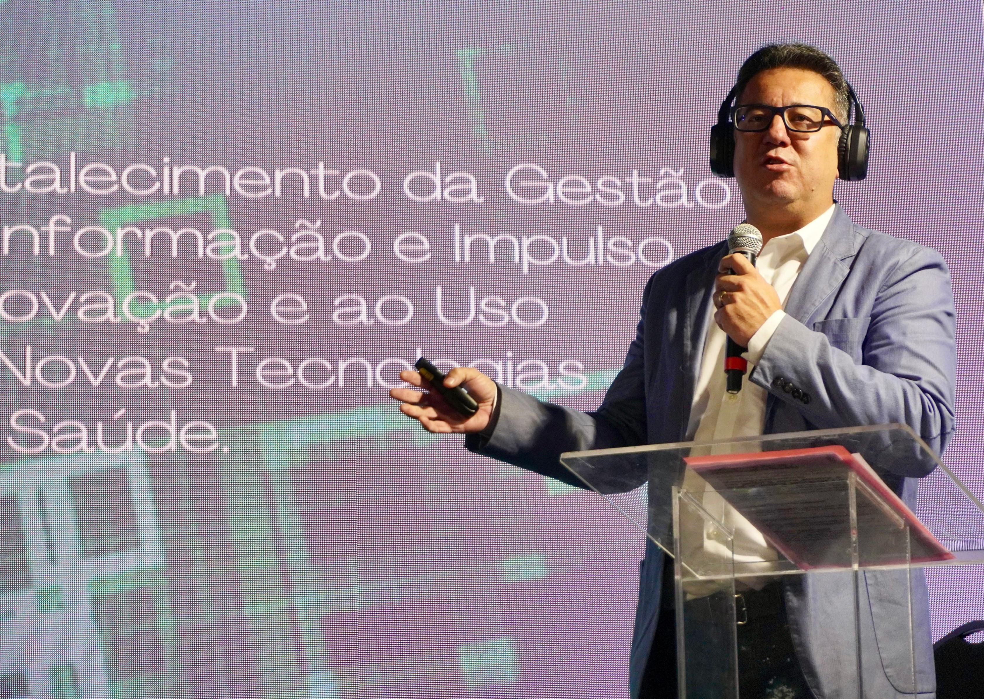 A imagem mostra um homem de meia-idade, caucasiano, em um palco. Ele usa um blazer azul claro sobre uma camisa social branca. Ele tem cabelo escuro e usa óculos e fones de ouvido pretos. Em uma das mãos, ele segura um microfone e na outra, um controle de apresentação, gesticulando enquanto fala. Ao fundo, há um telão com um texto em português que parece estar relacionado ao tema de sua fala, embora o texto esteja parcialmente ilegível