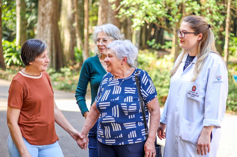 A imagem mostra quatro mulheres em um ambiente ao ar livre, provavelmente um parque, cercado por árvores. Três delas são idosas e estão em clima de conversa amigável. Uma mulher de cabelo curto, grisalho, veste camiseta marrom e calça clara; outra, de óculos e cabelos grisalhos soltos, usa blusa verde escura; e a terceira, também de cabelos brancos curtos, veste uma blusa azul estampada e segura a mão da mulher de camiseta marrom, sorrindo. Ao lado delas, está uma profissional de saúde jovem, usando jaleco branco com o brasão da Cidade de São Paulo – Secretaria Municipal da Saúde, acompanhando a interação. O clima da cena é de acolhimento, cuidado e bem-estar, transmitindo proximidade entre profissionais de saúde e a população idosa.