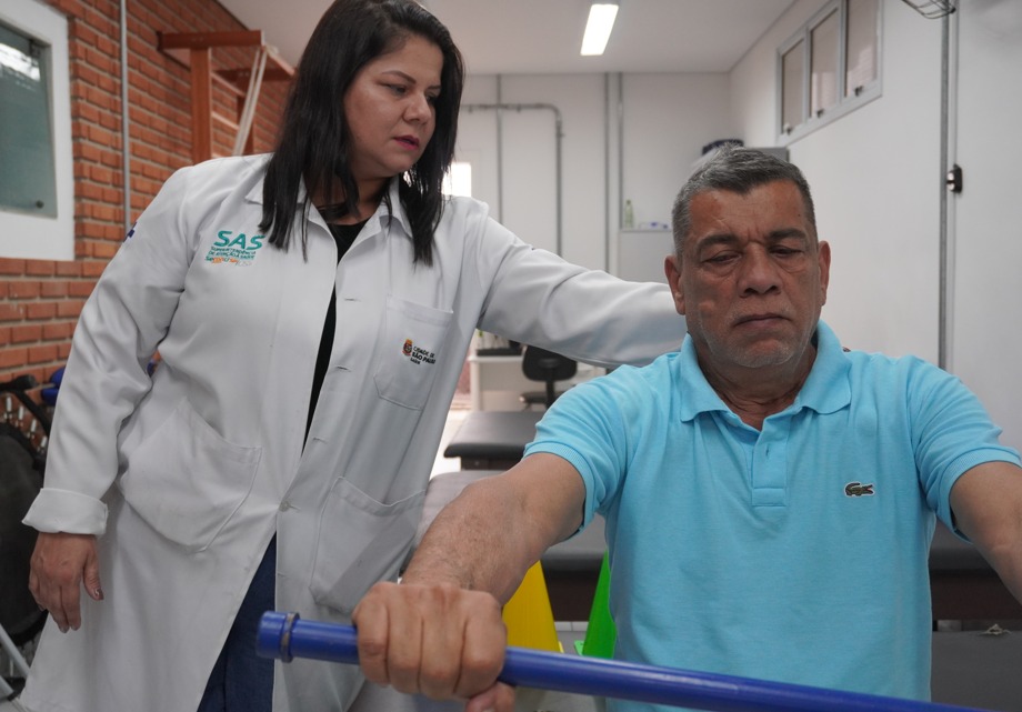 A imagem mostra um momento de atendimento de fisioterapia. Um homem, vestindo camisa polo azul, segura uma barra de exercício enquanto realiza um movimento orientado. Ao seu lado, uma profissional de saúde com jaleco branco e o logotipo da SAS – Secretaria Municipal da Saúde de São Paulo bordado no peito esquerdo acompanha de perto o exercício, apoiando e observando com atenção. O ambiente é uma sala de reabilitação, com paredes de tijolo aparente e equipamentos de fisioterapia visíveis ao fundo. A cena transmite cuidado, acompanhamento profissional e esforço do paciente em seu processo de recuperação.