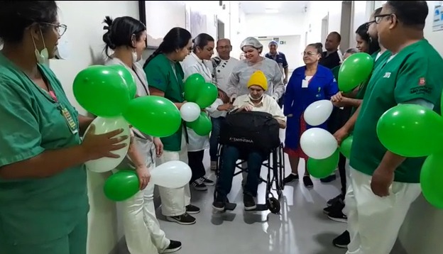 A imagem mostra um corredor de hospital onde um paciente, sentado em cadeira de rodas e usando gorro amarelo, é conduzido por profissionais de saúde. Ele está cercado por médicos, enfermeiros e funcionários vestidos com jalecos e uniformes, que comemoram sua saída segurando balões verdes e brancos. O clima é de celebração e acolhimento, transmitindo a ideia de alta hospitalar ou vitória após um período de internação.