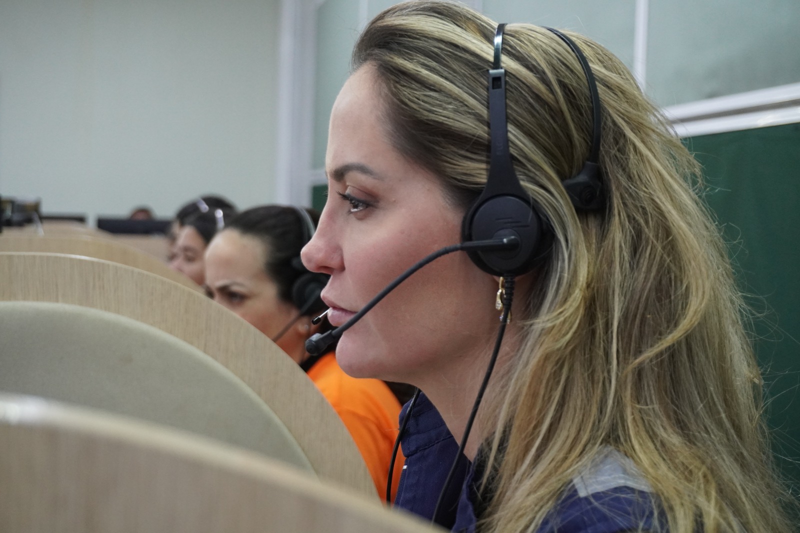 A foto mostra uma mulher jovem de cabelos loiros, em um local que parece uma central de atendimento; ela está de perfil, séria, e usa um headphone; ao seu lado, ao fundo, estão outras mulheres, parcialmente escondidas por divisórias, usando headphones