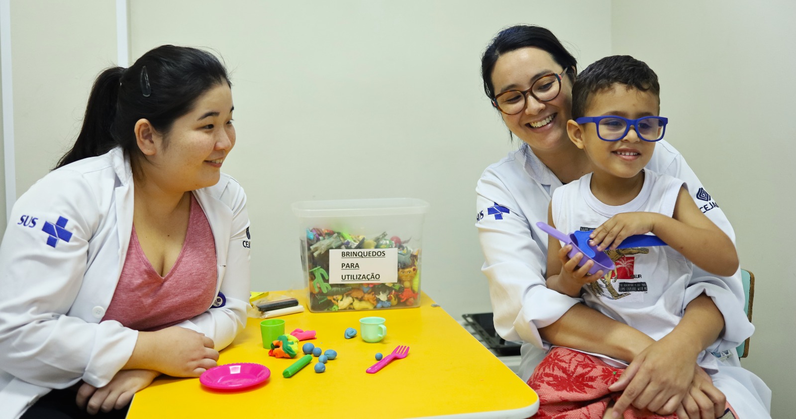 A imagem mostra um ambiente acolhedor de atendimento infantil em saúde. No centro, há uma mesa amarela com brinquedos de massinha, utensílios de plástico (pratinhos, talheres, xícara) e um pote transparente identificado com a placa “BRINQUEDOS PARA UTILIZAÇÃO”, cheio de pequenos bonecos e objetos coloridos. À esquerda, uma profissional de saúde com jaleco branco do SUS sorri enquanto observa a interação. À direita, outra profissional, também de jaleco, segura um menino pequeno de óculos azuis, que sorri enquanto brinca com uma peça de brinquedo roxa. Ambos parecem se divertir no momento.