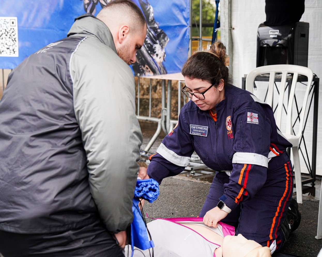 No chão, há um boneco de simulação usado para práticas de ressuscitação cardiopulmonar (RCP). Uma profissional do SAMU 192, identificada pelo uniforme azul-marinho com faixas refletivas e o crachá bordado com o nome “Enfermagem Giovanna Faciani”, está posicionada sobre o boneco, realizando compressões torácicas.  Ao lado dela, um homem de casaco cinza observa atentamente e segura uma bolsa de ventilação (ambú), demonstrando participação no exercício.