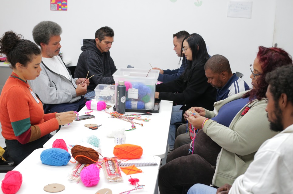 A imagem mostra um grupo de pessoas sentadas ao redor de uma mesa retangular branca, participando de uma oficina de artesanato com fios coloridos. Sobre a mesa estão espalhados novelos de lã de várias cores (rosa, azul, laranja, marrom), além de materiais de apoio como tesoura, papéis, caixas organizadoras e garrafa. Cada participante segura linhas ou agulhas de crochê, concentrado na atividade. O ambiente é simples e claro, com paredes brancas e algumas pequenas decorações fixadas nelas. A cena transmite um clima de aprendizado coletivo, foco e troca de experiências manuais.