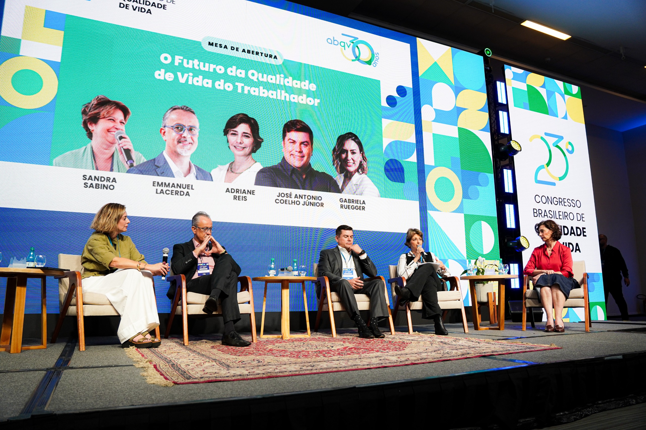 A imagem mostra uma mesa de abertura de um congresso, com cinco pessoas sentadas em cadeiras sobre um palco. Elas participam de um debate intitulado “O Futuro da Qualidade de Vida do Trabalhador”, conforme o grande painel projetado ao fundo. No painel, aparecem as fotos e nomes dos palestrantes: Sandra Sabino, Emmanuel Lacerda, Adriane Reis, José Antonio Coelho Junior e Gabriela Ruegger, além do logotipo do 23º Congresso Brasileiro de Qualidade de Vida, promovido pela ABQV (Associação Brasileira de Qualidade de Vida). O ambiente é bem iluminado e organizado, com um tapete sobre o palco, mesas laterais com garrafas d’água e um grande painel colorido em tons de azul, verde e amarelo, reforçando a identidade visual do evento. À direita, também é possível ver o logotipo do congresso projetado verticalmente em um telão.