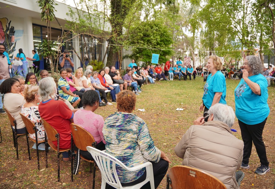 A imagem mostra um grupo grande de pessoas reunidas em um espaço ao ar livre, provavelmente um jardim ou pátio, cercado por árvores e vegetação. A cena tem um clima de convivência e alegria. As pessoas estão sentadas em cadeiras dispostas em formato circular, participando de uma atividade coletiva. A maioria dos participantes são idosos, e duas mulheres em pé, vestindo camisetas azuis, parecem conduzir a dinâmica ou conversar com o grupo. Ao fundo, vê-se mais pessoas observando ou participando, além de um prédio com grandes janelas e murais coloridos, que pode ser uma unidade de saúde ou centro comunitário. O ambiente transmite integração, cuidado e convivência social entre os participantes.