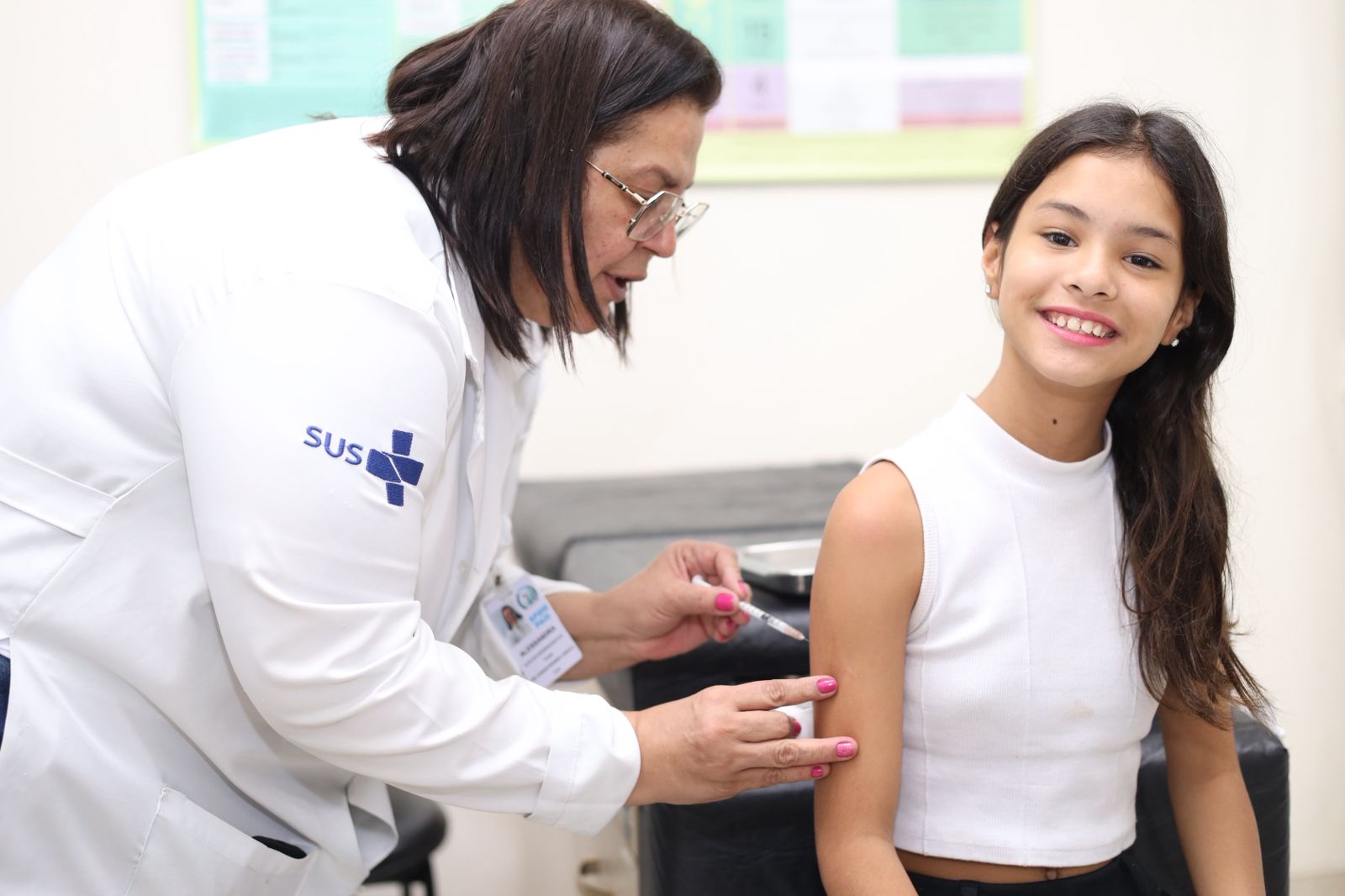 A imagem mostra uma profissional de saúde do SUS aplicando uma vacina em uma menina. A profissional, vestida com jaleco branco e crachá de identificação, segura cuidadosamente uma seringa enquanto realiza o procedimento no braço esquerdo da criança. A menina, com blusa branca sem mangas, sorri para a câmera, transmitindo tranquilidade e confiança. O ambiente é o de uma unidade de saúde, com paredes claras e cartazes informativos ao fundo. A cena representa um momento de cuidado e prevenção em saúde pública, destacando a importância da vacinação infantil.