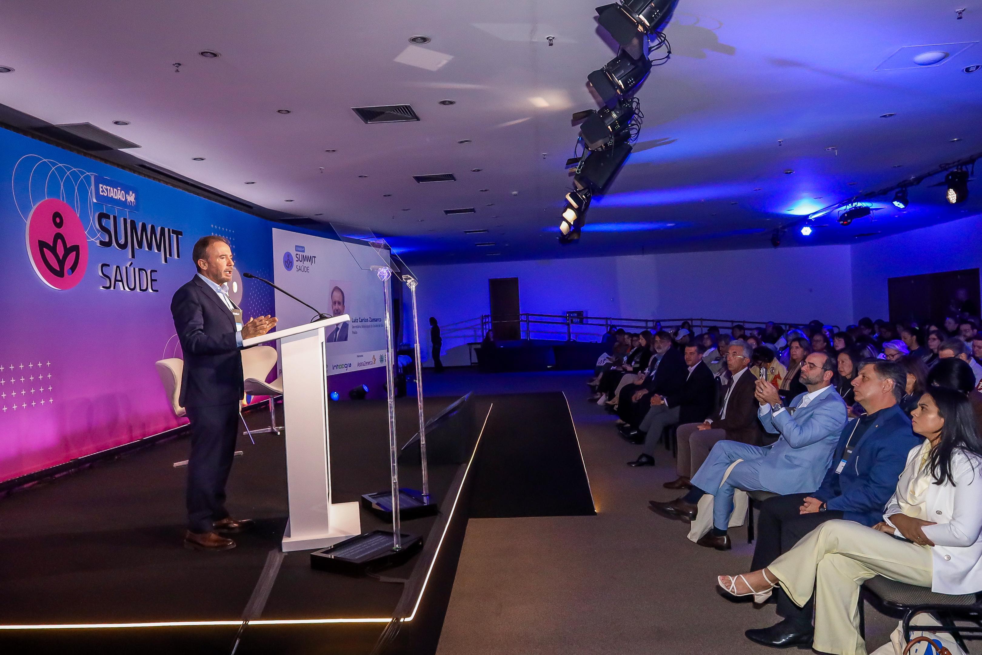 A imagem mostra um evento corporativo ou congresso de saúde, identificado pelo painel no palco com o logotipo “Summit Saúde – Estadão”. Um palestrante, em traje formal e posicionado atrás de um púlpito branco, fala ao microfone diante de uma plateia numerosa e atenta. O ambiente é um auditório moderno, com iluminação azul e roxa, equipamentos de som e refletores suspensos no teto. Ao fundo, vê-se uma tela projetando a identidade visual do evento e o nome do palestrante. As pessoas sentadas nas primeiras fileiras — também de terno e roupas sociais — acompanham a fala, algumas registrando o momento com o celular.