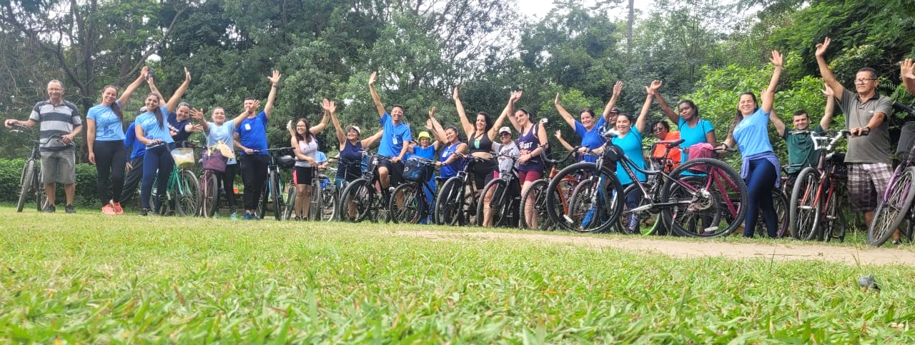 A foto mostra um grupo animado de pessoas reunidas ao ar livre para uma atividade ciclística. Homens e mulheres de diferentes idades posam lado a lado com suas bicicletas, formando uma linha e erguendo os braços em sinal de alegria e celebração. Muitos vestem camisetas esportivas, especialmente em tons de azul, e sorriem para a câmera. O cenário é natural, com gramado verde em primeiro plano e árvores ao fundo, indicando um parque ou área de trilha. A imagem transmite um clima de descontração, companheirismo e incentivo à prática de atividades físicas ao ar livre.