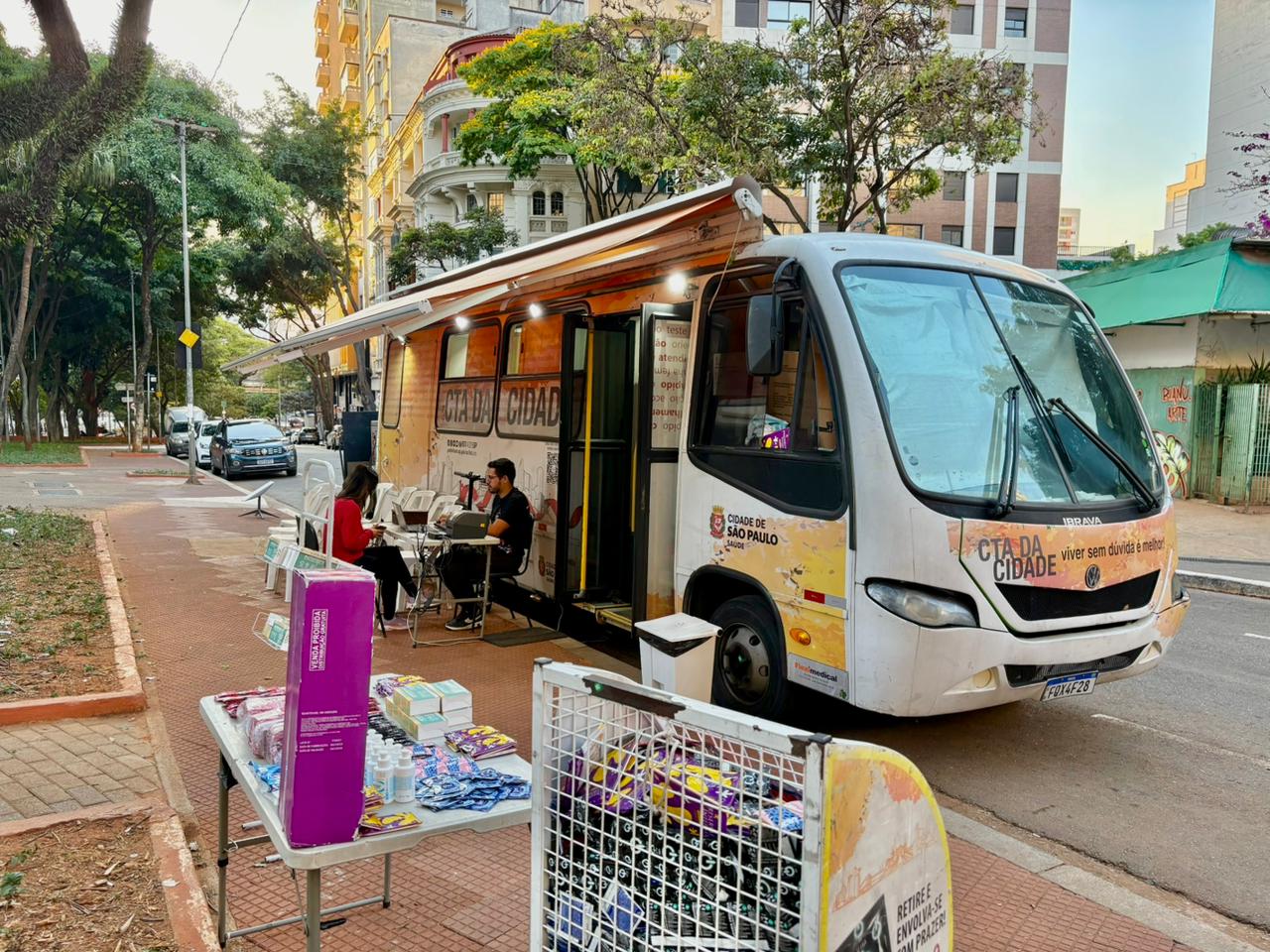 A imagem mostra uma unidade móvel de saúde estacionada em uma calçada arborizada, em uma área urbana. Trata-se de um ônibus identificado como CTA da Cidade, da Prefeitura de São Paulo, adaptado para atendimento em saúde. O veículo está com o toldo lateral aberto, formando uma área coberta onde há cadeiras e uma pequena mesa. Duas pessoas estão sentadas, sendo atendidas por um profissional, em um ambiente que sugere acolhimento e orientação em saúde. Em primeiro plano, há uma mesa com diversos insumos organizados, como preservativos, materiais informativos e caixas de produtos voltados à prevenção de infecções sexualmente transmissíveis. Ao lado, uma estrutura metálica contém mais materiais embalados. Ao fundo, aparecem prédios residenciais, árvores e carros estacionados na rua, indicando que o atendimento ocorre em via pública, integrado ao cotidiano da cidade. A cena transmite a ideia de um serviço itinerante, acessível e voltado à promoção da saúde e da prevenção.
