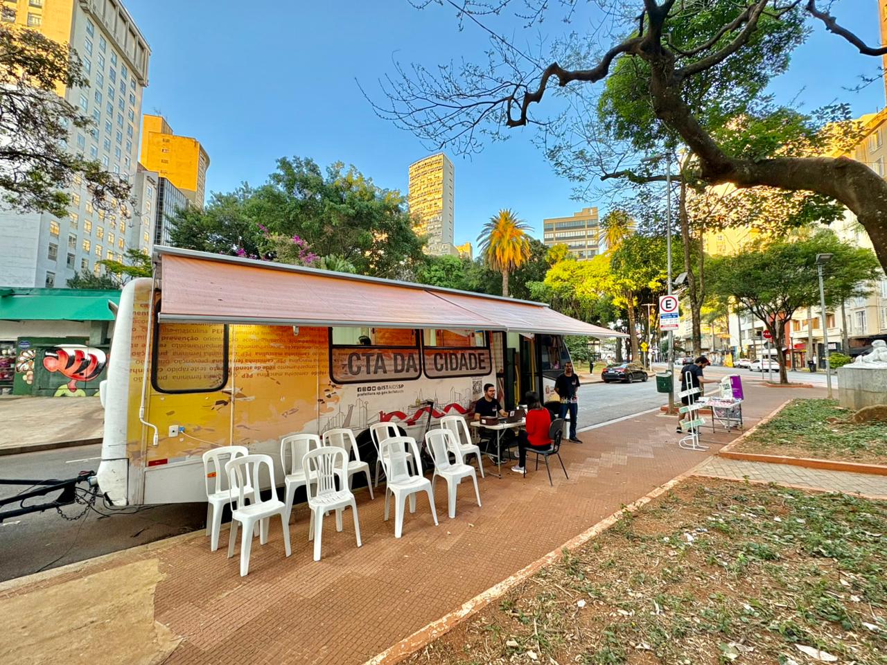 A imagem mostra uma unidade móvel de atendimento instalada em uma área urbana arborizada, provavelmente em uma praça ou canteiro central de uma avenida. Trata-se de um trailer adaptado, com uma cobertura lateral estendida, onde há cadeiras plásticas organizadas para espera. Na lateral do veículo, lê-se “CTA da Cidade”, indicando que pode ser um serviço de saúde pública voltado para testagem e aconselhamento (como HIV, ISTs ou outras ações de prevenção). Algumas pessoas estão sentadas sendo atendidas por profissionais, enquanto outras aguardam. Há mesas simples com cadeiras, criando um ambiente de atendimento informal e acessível. Ao redor, vê-se um cenário típico de centro urbano, com prédios altos, árvores grandes e uma rua com carros estacionados. A presença de sinalização de trânsito e ciclovia reforça que é um espaço público movimentado.