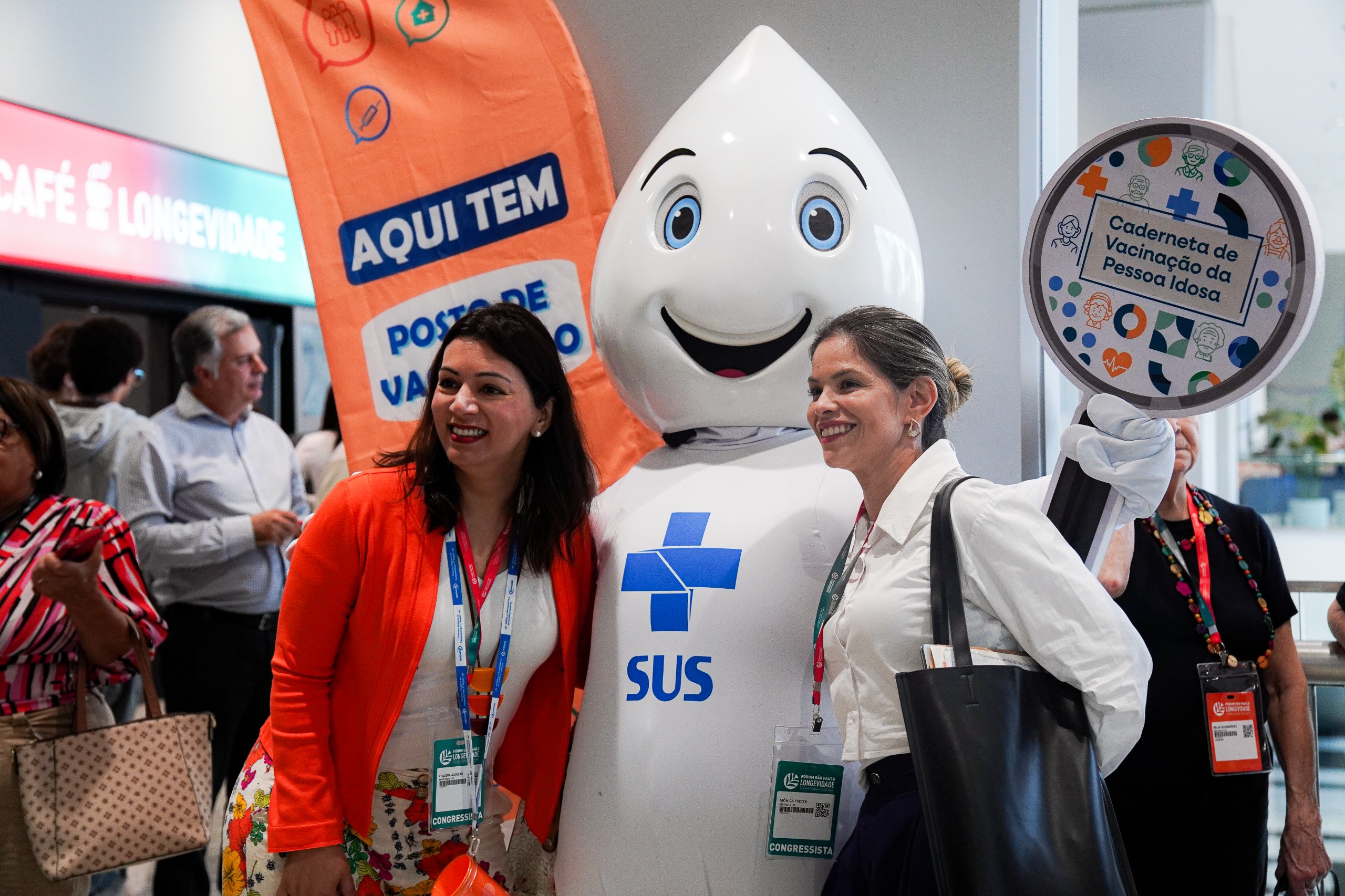 A imagem mostra um ambiente de evento, com várias pessoas sorrindo e interagindo. No centro, aparece o personagem Zé Gotinha, mascote das campanhas de vacinação do SUS (Sistema Único de Saúde), posando para fotos com duas mulheres identificadas como congressistas por seus crachás. Uma delas veste casaco laranja e a outra, camisa branca.  Ao fundo, há um banner laranja com os dizeres “AQUI TEM POSTO DE VACINAÇÃO”, e o mascote segura um grande símbolo circular com o texto “Caderneta de Vacinação da Pessoa Idosa”. Na lateral esquerda da foto, vê-se uma porta encimada por uma placa onde lê-se 