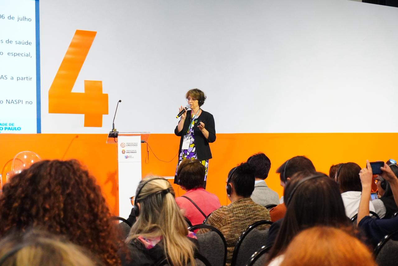 A imagem mostra uma mulher falando ao microfone em um auditório, diante de um público sentado. Ela está em pé, próxima a um púlpito com o logotipo da Prefeitura de São Paulo e do Fórum São Paulo Longevidade. No fundo, há um grande painel branco e laranja com o número 4 destacado. O público, composto por várias pessoas, parece atento à apresentação, e alguns usam fones de ouvido. O ambiente é iluminado e tem aspecto profissional, típico de um evento ou congresso.