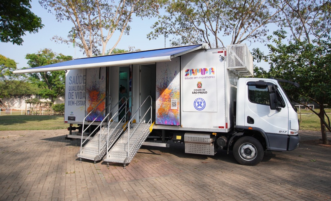 A imagem mostra uma unidade móvel de saúde da Prefeitura de São Paulo estacionada em um ambiente externo arborizado. O veículo é um caminhão adaptado com um toldo azul e duas entradas acessíveis por escadas metálicas. Na lateral do caminhão, estão estampadas as palavras 