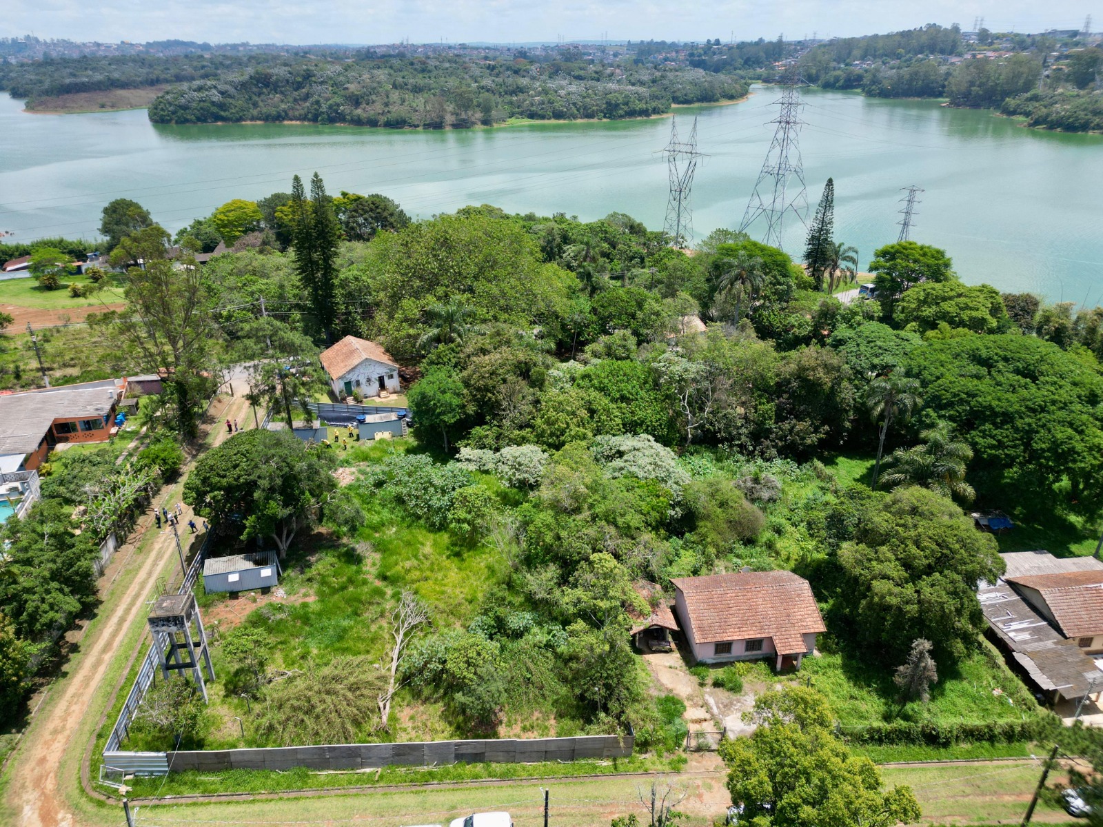 A imagem mostra uma vista aérea de uma área verde e arborizada às margens de uma represa. Entre as árvores, é possível ver algumas construções de pequeno porte, com telhados de cerâmica, cercadas por vegetação densa. Há também uma via de terra que atravessa parte do terreno e algumas pessoas caminhando por ela. Ao fundo, nota-se a ampla extensão da represa Billings, com torres de transmissão de energia cruzando a paisagem. O cenário tem aspecto rural, com bastante natureza e poucas edificações, sugerindo um ambiente tranquilo e afastado da área urbana.
