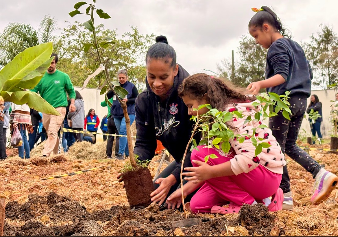 A imagem mostra um grupo de pessoas participando de uma atividade de plantio ao ar livre, em um terreno de terra marrom e fofa. Em primeiro plano, uma mulher de pele negra e cabelo preso em coque ajuda uma menina pequena, de cabelos castanhos cacheados, vestida com moletom claro estampado e calça rosa, a plantar uma muda de árvore. Ambas estão agachadas, concentradas na tarefa, segurando a muda com cuidado. Ao fundo, outras pessoas — homens e mulheres — também participam da ação, algumas com roupas de frio e jaquetas, indicando clima fresco. O céu está nublado e o ambiente tem árvores e vegetação ao redor