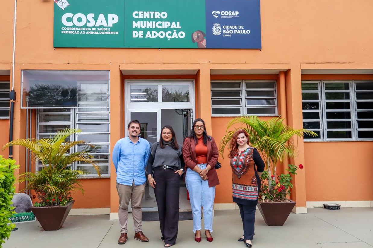A imagem mostra quatro pessoas posando para uma foto em frente ao prédio do Centro Municipal de Adoção, da Coordenadoria de Saúde e Proteção ao Animal Doméstico (COSAP), da Prefeitura de São Paulo. Elas estão sorrindo e posicionadas lado a lado, diante da fachada pintada em tom alaranjado, que traz placas verdes e azuis com o nome da instituição. Há vasos com plantas ao redor da entrada, contribuindo para um ambiente acolhedor e bem cuidado.
