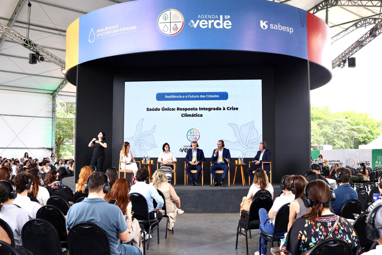 A imagem mostra um evento realizado em um grande auditório coberto, com público atento e estrutura profissional. No palco, há cinco pessoas sentadas — três homens e duas mulheres — participando de um painel de debate. Uma intérprete de Libras está posicionada à esquerda, traduzindo as falas dos participantes.  No telão central atrás do grupo, lê-se o tema da mesa: “Saúde Única: Resposta Integrada à Crise Climática”, dentro do eixo “Resiliência e o Futuro das Cidades”, que faz parte da Agenda SP +Verde, iniciativa da Sabesp.  A plateia, composta por dezenas de pessoas, acompanha a discussão usando fones de ouvido. O ambiente é iluminado, com vegetação visível ao fundo, e o clima geral é de um encontro técnico e institucional voltado à sustentabilidade e saúde pública.