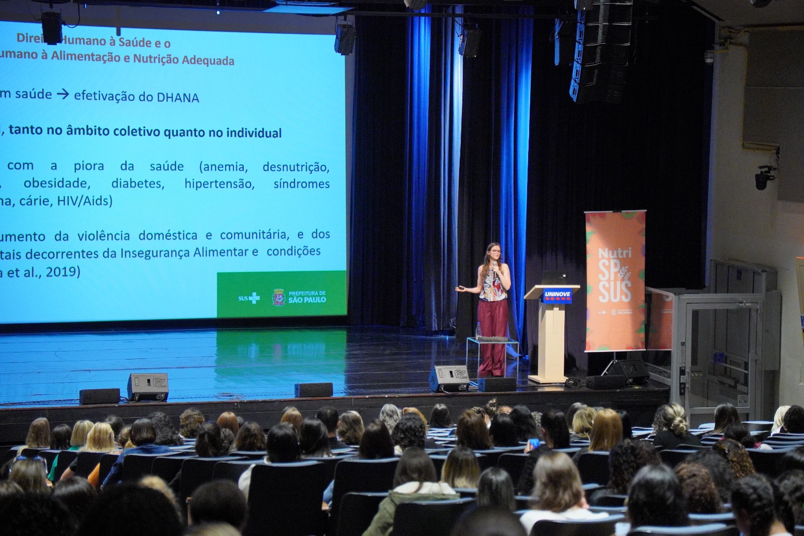 A imagem mostra um auditório cheio, com várias pessoas sentadas assistindo a uma palestra. No palco, há uma mulher em pé, falando ao microfone e gesticulando, ao lado de um púlpito com o logotipo da Uninove. Atrás dela, um grande telão exibe um slide de apresentação sobre Direito Humano à Saúde e à Alimentação e Nutrição Adequada, com os logotipos do SUS e da Prefeitura de São Paulo. Também há um banner lateral com a identidade visual do evento “Nutri SP + SUS”. O ambiente é bem iluminado, com cortinas azuis e estrutura de som, e o público parece atento à exposição.