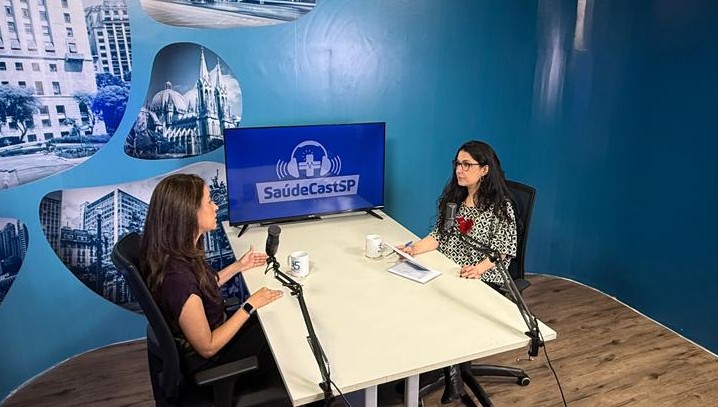 A imagem mostra duas mulheres sentadas frente a frente em um estúdio de gravação, participando de uma conversa ou entrevista em formato de podcast. Elas estão posicionadas em uma mesa branca, cada uma com um microfone profissional à sua frente, indicando um ambiente preparado para captação de áudio. Ao fundo, há um painel azul com o logotipo “SaúdeCastSP” exibido em uma tela, além de imagens urbanas que remetem à cidade de São Paulo. O espaço é bem iluminado, organizado e com aparência institucional, sugerindo um programa voltado à área da saúde ou a temas relacionados a políticas públicas e serviços. Uma das participantes parece conduzir a conversa, gesticulando enquanto fala, enquanto a outra escuta atentamente, com anotações sobre a mesa. O cenário transmite profissionalismo e um clima de diálogo informativo.
