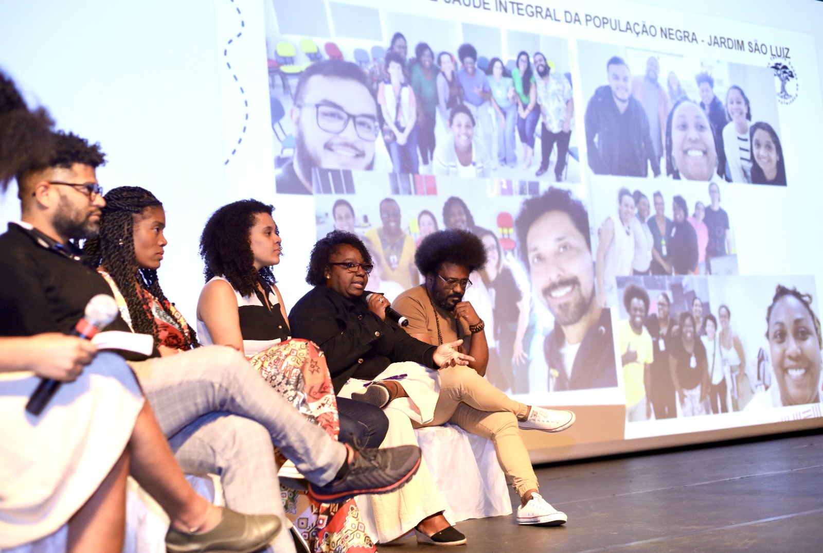 A imagem mostra uma mesa de debate em um auditório, com cinco pessoas sentadas em cadeiras no palco. Todas parecem participar de uma conversa pública ou conferência. Uma das mulheres ao centro segura um microfone enquanto fala, e as demais pessoas observam atentamente. À esquerda, uma pessoa segura outro microfone e um papel.  O grupo é diverso, com homens e mulheres, todos vestidos de maneira casual e confortável. No fundo do palco, um telão exibe uma grande montagem de fotos com várias pessoas sorrindo, provavelmente integrantes de algum projeto ou equipe. No topo da projeção é possível ler parte de um texto que menciona “Saúde Integral da População Negra – Jardim São Luiz”.