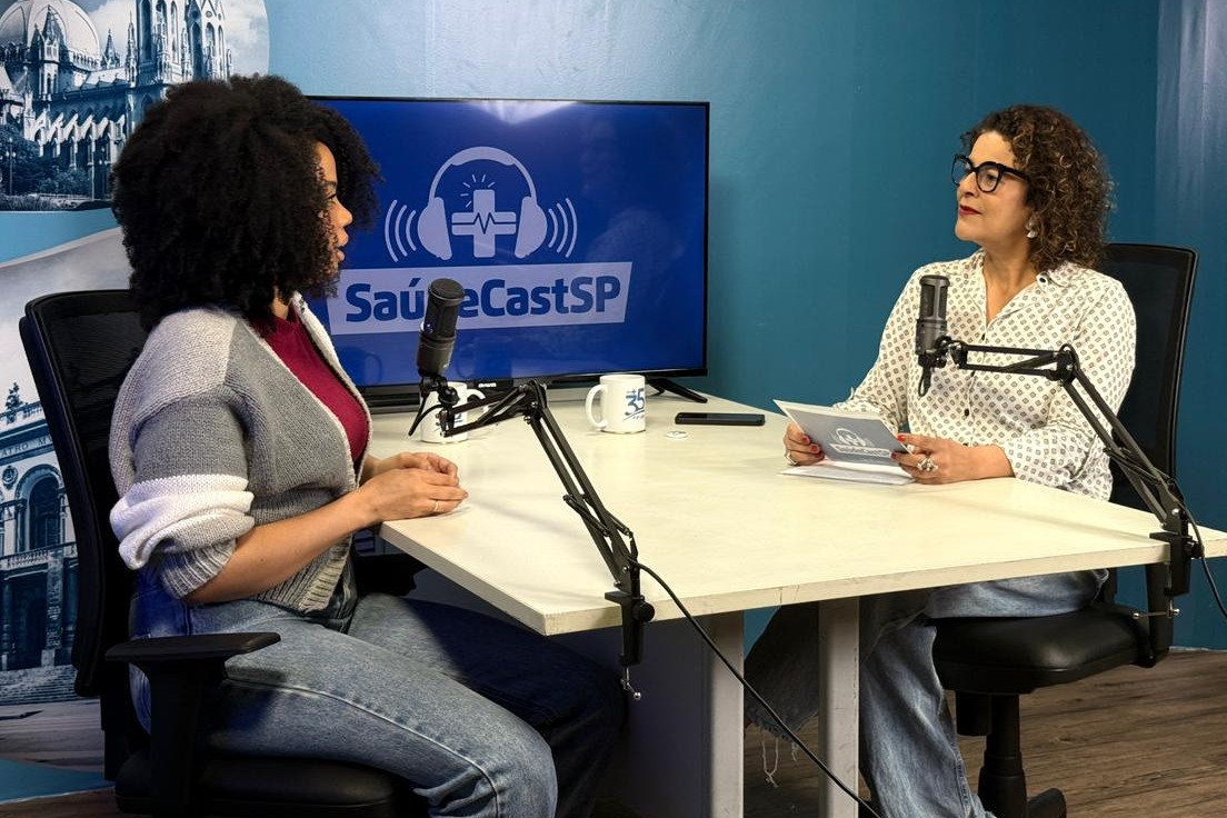 A imagem retrata um cenário de gravação de um podcast ou entrevista em estúdio, com foco em duas mulheres sentadas à mesa, cada uma com seu microfone. À Esquerda está Uma mulher jovem, de pele escura e cabelos crespos volumosos. Ela veste uma blusa escura (possivelmente vinho ou bordô) sob um cardigã cinza claro com detalhes brancos nas mangas. Ela está olhando para a mulher à direita, em posição de escuta ou resposta. À direita está uma mulher de pele clara, com cabelos cacheados curtos e grisalhos. Ela veste uma camisa social de cor clara (branca ou bege) com estampa escura (geométrica ou de bolinhas) e óculos de aros pretos. Ela segura alguns papéis ou um livreto (talvez um roteiro) e olha diretamente para a mulher à esquerda, em posição de condução da entrevista ou diálogo.