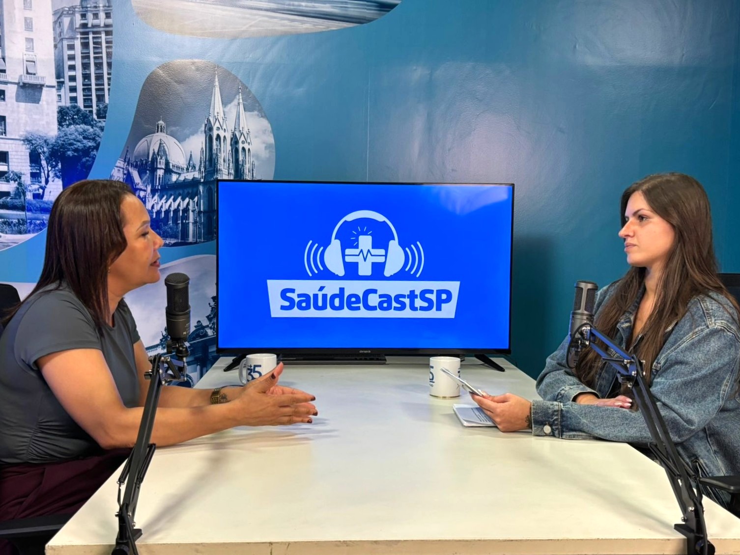 A imagem mostra duas mulheres sentadas frente a frente em um estúdio de gravação de podcast ou entrevista. Entre elas, há uma mesa clara com dois microfones profissionais montados em suportes articulados e duas canecas. Ao fundo, uma televisão exibe o logotipo “SaúdeCastSP”, com um símbolo de fone de ouvido e uma cruz, indicando que se trata de um programa voltado à área da saúde. A parede tem tons de azul e traz uma imagem urbana ao fundo. À esquerda, uma mulher fala enquanto gesticula com as mãos, demonstrando envolvimento na conversa. À direita, a outra mulher escuta atentamente, segurando um celular ou bloco de notas, como se estivesse acompanhando perguntas ou anotações.