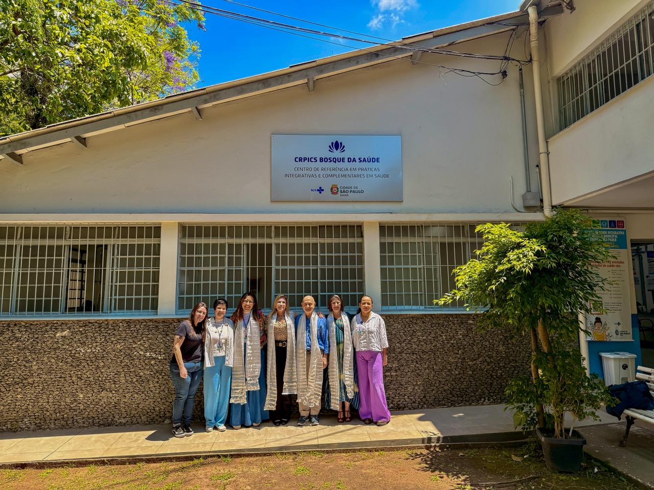 A imagem mostra a fachada do Centro de Referência em Práticas Integrativas e Complementares em Saúde (CRPICS) Bosque da Saúde, na região sudeste de São Paulo. Na frente do prédio, que é claro e possui janelas com grades, há um grupo de sete pessoas posando para a foto. Elas estão lado a lado, sorrindo, em um dia ensolarado. Algumas usam roupas profissionais e crachás; três delas vestem estolas claras com padrões verticais, sugerindo algum tipo de traje institucional ou cerimonial. À direita da imagem existe uma planta em vaso, e ao fundo é possível ver placas informativas do serviço de saúde.