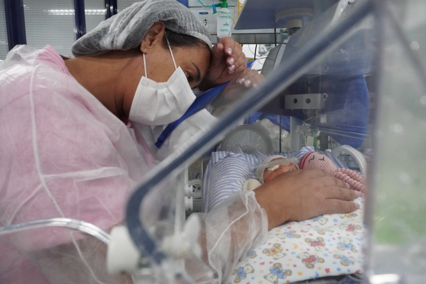 Na foto, uma mulher adulta, está inclinada sobre uma incubadora de bebê. Ela usa uma touca cirúrgica cinza clara e uma máscara facial branca que cobre seu nariz e boca. Sua pele parece ter um tom moreno claro, e seus cabelos escuros estão completamente cobertos pela touca. Seus olhos estão voltados para o bebê dentro da incubadora. Uma de suas mãos está delicadamente posicionada sobre a lateral da incubadora, tocando a bebê, enquanto a outra parece estar perto de seu rosto. Ela veste uma espécie de camisola ou roupão hospitalar de cor rosa claro.  Dentro da incubadora, podemos ver um bebê muito pequeno, deitado de costas sobre um tecido estampado com desenhos infantis coloridos. O bebê parece estar envolto em um tecido claro e tem alguns tubos ou fios finos conectados ao seu corpo, sugerindo que ele está recebendo cuidados médicos.