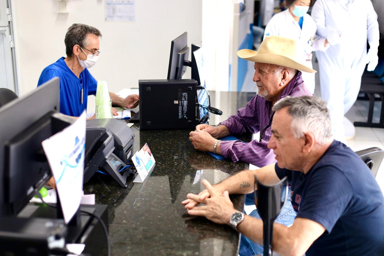 A imagem mostra um ambiente que parece ser a recepção de uma unidade de saúde ou clínica. Três homens estão sentados no balcão de atendimento, sendo atendidos por um profissional usando máscara e roupa azul.
