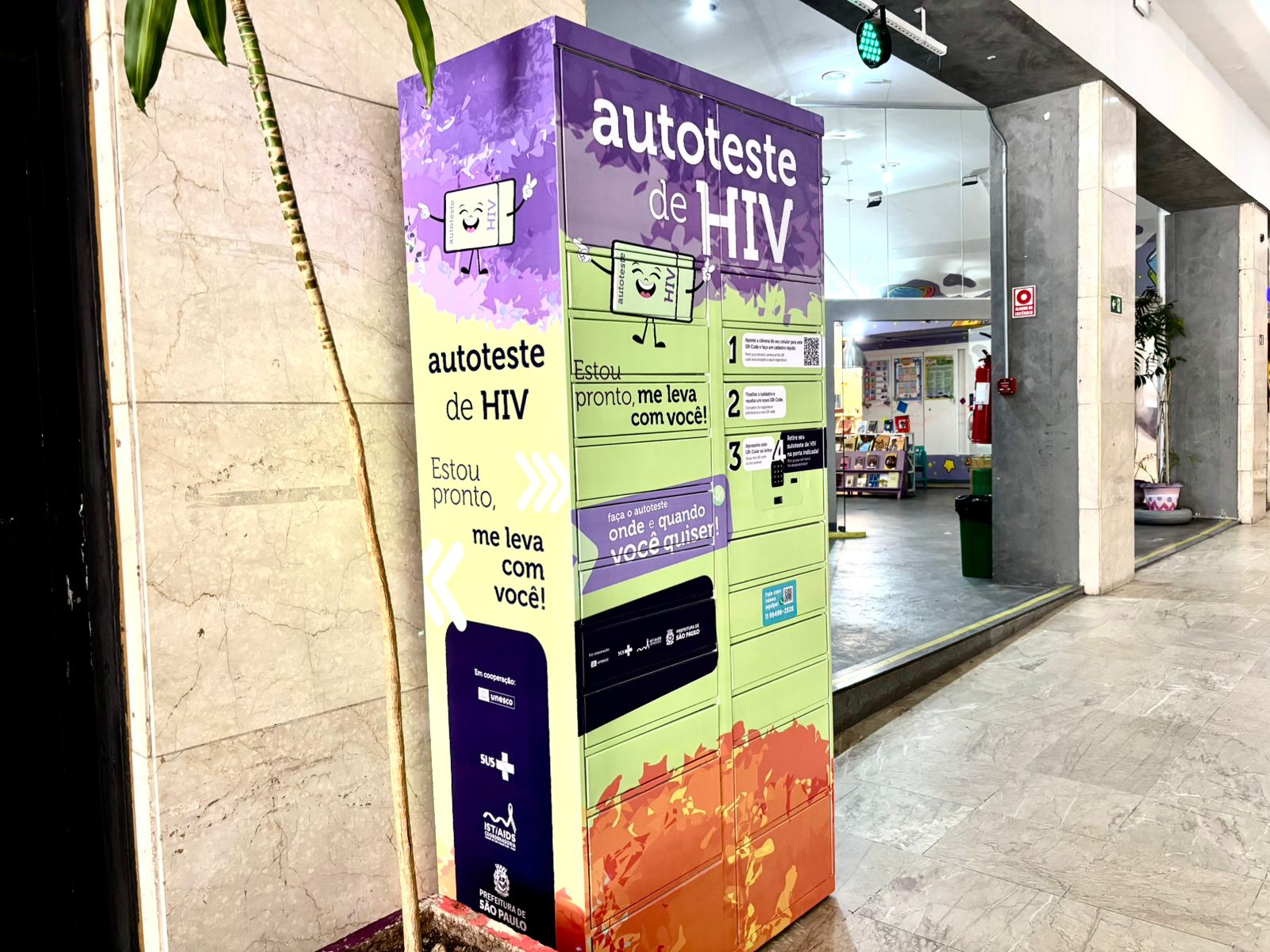 A imagem mostra uma máquina de distribuição de autotestes de HIV instalada em um corredor interno. O equipamento é colorido, com tons de roxo, verde e laranja, e exibe mensagens como “autoteste de HIV”, “Estou pronto, me leva com você!” e instruções ilustradas sobre como retirar e utilizar o teste. Há ícones e setas chamando a atenção para o serviço, além de logotipos de instituições públicas. Ao fundo, é possível ver um espaço interno com iluminação clara, paredes brancas, colunas e uma área que parece ser um ambiente de atendimento ou exposição, com prateleiras e itens coloridos. Uma planta alta aparece à esquerda da máquina.