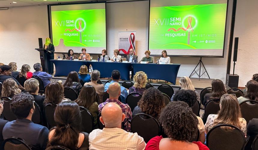 A imagem mostra um auditório cheio, com dezenas de pessoas sentadas e voltadas para uma mesa composta por sete palestrantes. Eles estão posicionados à frente da sala, atrás de uma mesa longa azul, participando do XVII Seminário de Pesquisas em IST/Aids, conforme indicado nos dois telões projetados atrás deles, que exibem o nome do evento em um fundo verde vibrante.  À esquerda, uma mulher de vestido escuro está em um púlpito, aparentemente conduzindo ou apresentando a mesa. Ao lado dos telões, há um banner do programa municipal de IST/Aids, com o laço vermelho característico.  O público é diverso, com pessoas de diferentes idades e estilos, todas atentas às falas. O ambiente é organizado, com cadeiras alinhadas e iluminação clara, típico de eventos técnicos ou científicos.
