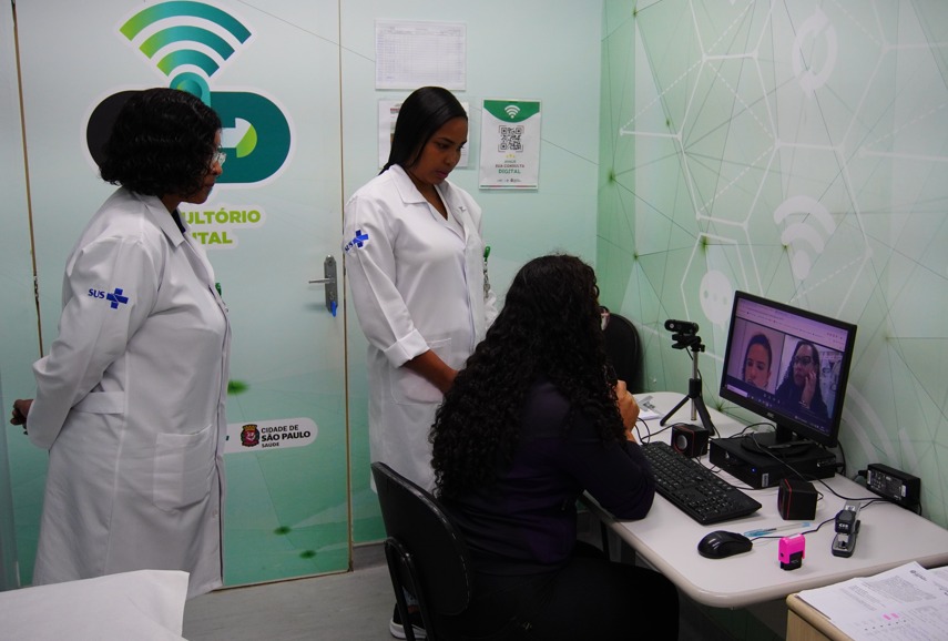 A imagem mostra uma sala de teleatendimento em uma unidade de saúde. Três profissionais estão presentes: duas delas vestem jalecos brancos com o logo do SUS no braço, e a terceira está sentada à frente de um computador, conduzindo uma videochamada.  Na tela do monitor aparecem duas pessoas participando da teleconsulta. Sobre a mesa, há equipamentos como teclado, mouse, câmera externa posicionada em um tripé pequeno, caixas de som e documentos. Ao fundo, a parede tem um padrão gráfico moderno, com ícones de conexão, reforçando o tema digital. A porta ao lado exibe o logotipo do Consultório Digital da Prefeitura de São Paulo.  O ambiente transmite a ideia de atendimento remoto, tecnologia aplicada à saúde e trabalho colaborativo entre profissionais.