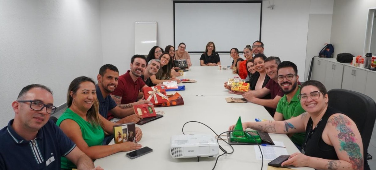 A imagem mostra um grupo de cerca de 20 pessoas reunidas em uma sala de reunião, sentadas ao redor de uma mesa longa e retangular. Todas parecem à vontade e sorrindo para a câmera, o que transmite um clima descontraído e colaborativo.  Sobre a mesa, há caixas de presentes, doces e lembranças, além de celulares, cadernos e um projetor, sugerindo um encontro que pode ter caráter de reunião de trabalho com momento de confraternização, como uma comemoração interna ou encerramento de atividade. No centro da mesa, o projetor está ligado, apontando para uma tela branca ao fundo da sala.  O ambiente é simples e funcional, com paredes claras, armários laterais e iluminação uniforme, típico de um espaço institucional ou corporativo. As pessoas vestem roupas casuais, algumas em tons semelhantes, reforçando a sensação de integração e equipe.