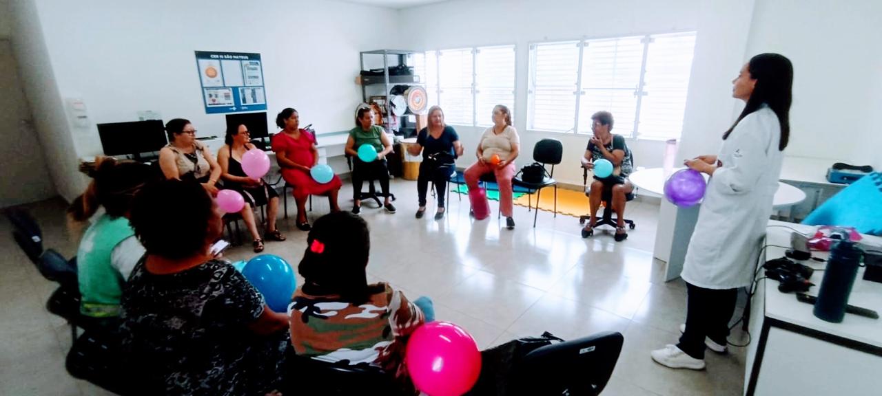 A imagem mostra uma roda de conversa do grupo de apoio aos cuidadores da CER São Mateus. As participantes — em sua maioria mulheres — estão sentadas em círculo dentro de uma sala ampla e iluminada, segurando balões coloridos usados como parte das dinâmicas do encontro. À direita, uma profissional de saúde, de jaleco branco, conduz a atividade e interage com o grupo. O clima é de acolhimento e participação ativa, em um ambiente que reforça a troca de experiências, o apoio emocional e o cuidado com quem cuida.