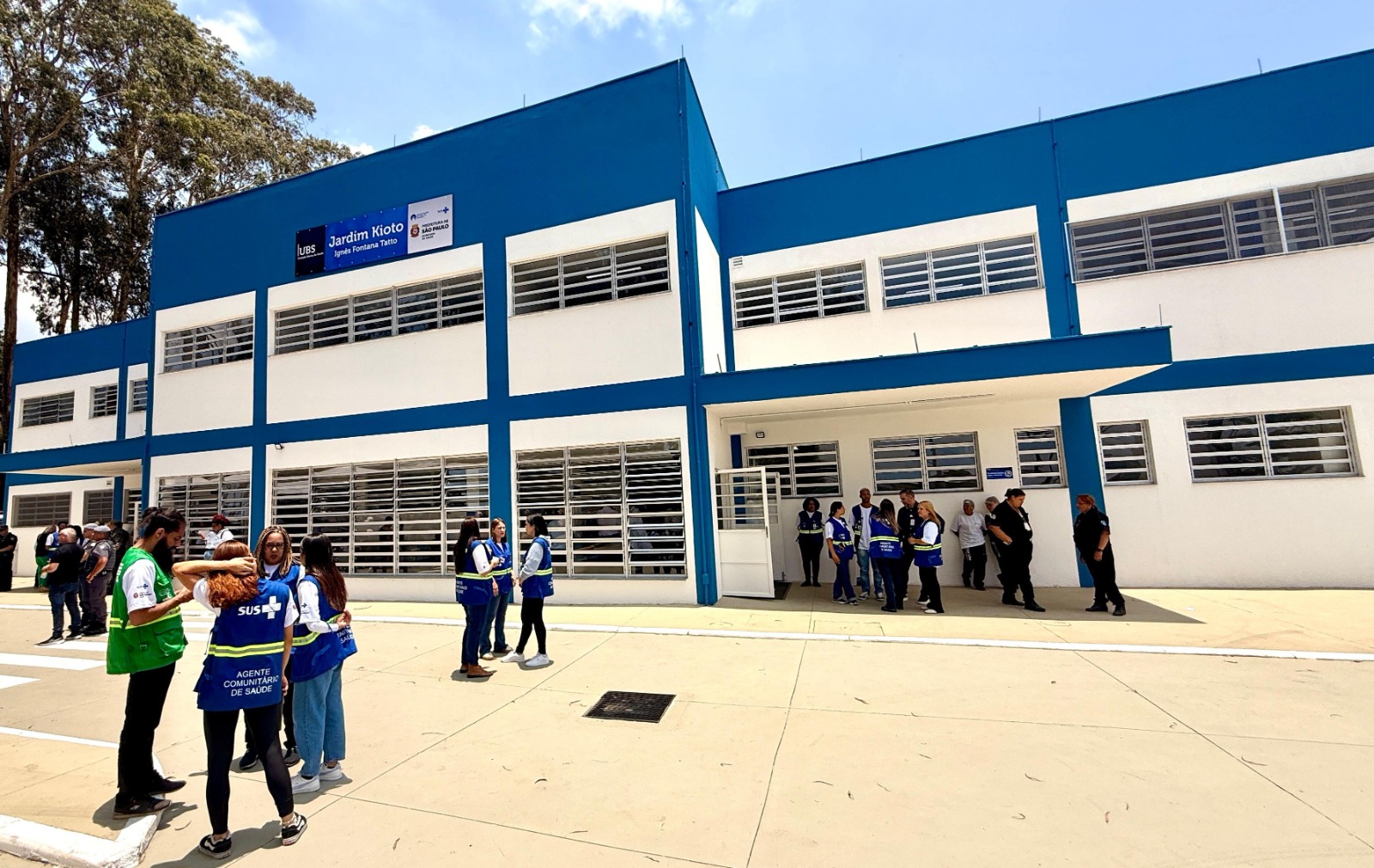 A imagem mostra a fachada de uma Unidade Básica de Saúde recém-inaugurada, identificada como UBS Jardim Kioto – Ignês Fontana Tatto. O prédio é amplo, de dois andares, com pintura branca e detalhes em azul, além de muitas janelas alinhadas horizontalmente. Em frente à unidade, diversos profissionais de saúde — alguns usando coletes do SUS e identificados como agentes comunitários de saúde — conversam em pequenos grupos. Outras pessoas também estão reunidas próximo à entrada, dando ao ambiente um clima de movimentação típica de evento de abertura ou visita técnica. Ao fundo, árvores altas completam a paisagem em um dia claro e ensolarado.