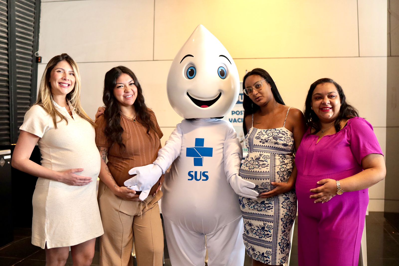 A imagem mostra quatro mulheres grávidas posando para a foto ao lado do mascote do SUS, o Zé Gotinha. Todas estão sorrindo ou demonstrando expressão amistosa, com as mãos apoiadas nas próprias barrigas, em um gesto típico de gestantes. O Zé Gotinha está no centro, com sua roupa branca característica, símbolo azul do SUS no peito e um grande sorriso no rosto. As mulheres vestem roupas confortáveis em tons claros e vibrantes — bege, marrom, azul estampado e rosa. O ambiente parece ser interno, bem iluminado, provavelmente relacionado a uma ação de saúde ou vacinação, já que há um cartaz do SUS ao fundo.